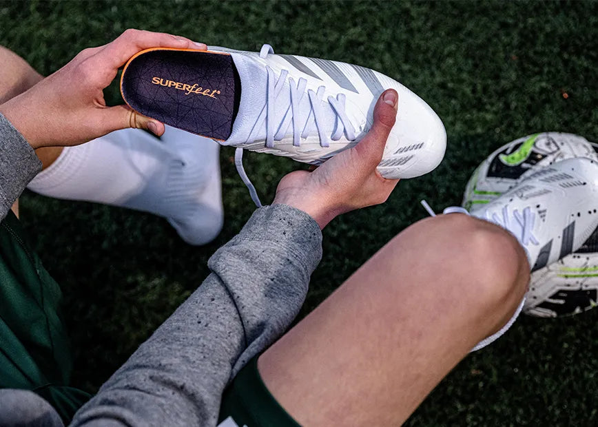 Man sitting on soccer turf inserting Superfeet All-Purpose Cushion Insoles into his soccer cleats.