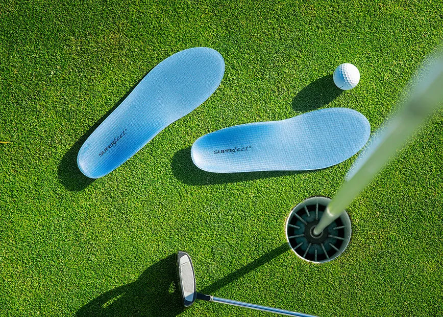 A pair of Superfeet All-Purpose Support Medium Arch insoles sitting on a golf green next to a golf ball, golf club, and hole. 