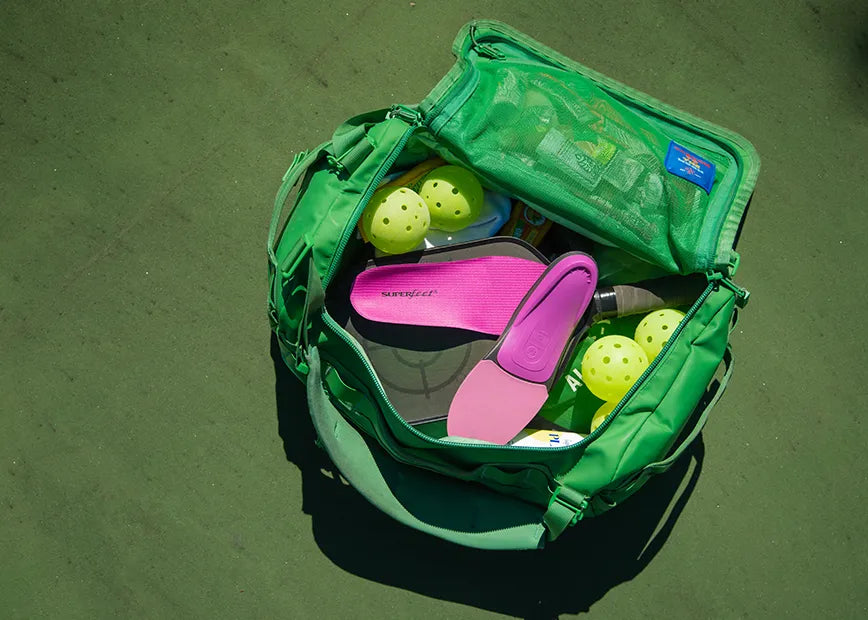 Gym bag sitting on a pickleball court with pickleball racquets, balls, and Superfeet All-Purpose Women's High Impact Support insoles inside.