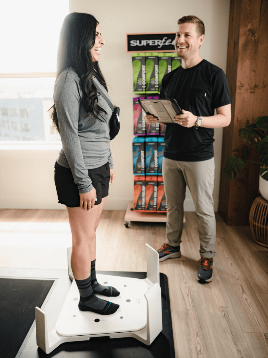 A shopper standing on a white rectagular 3D foot scanning platform, being assisted by a sales associate holding a smart tablet