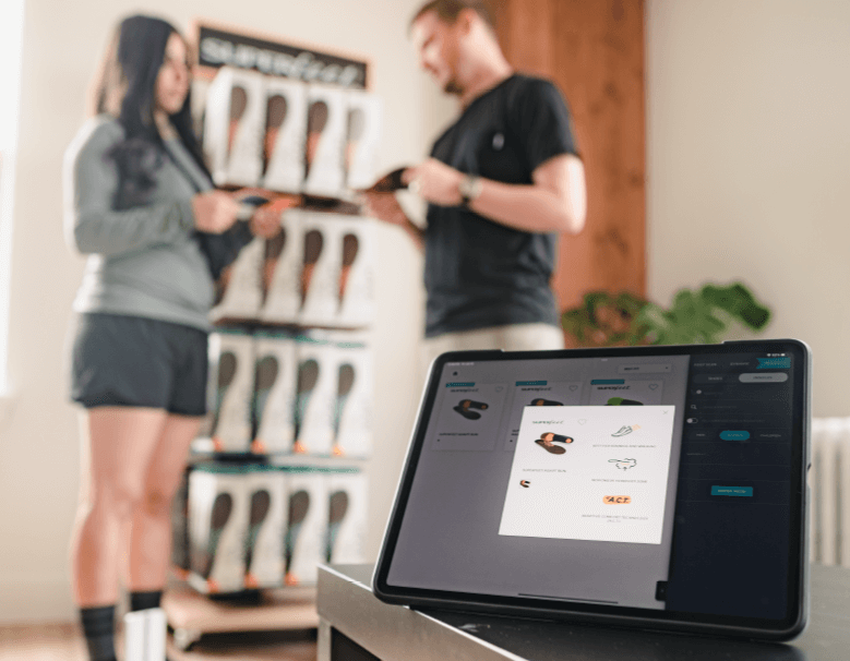 Your Best Superfeet - A smart tablet stands on a bench top in the foreground with a Superfeet trim-to-fit insole recommendation based on the 3D scan and gait analysis. In the background, a sales associate shows the recommended insole to the shopper